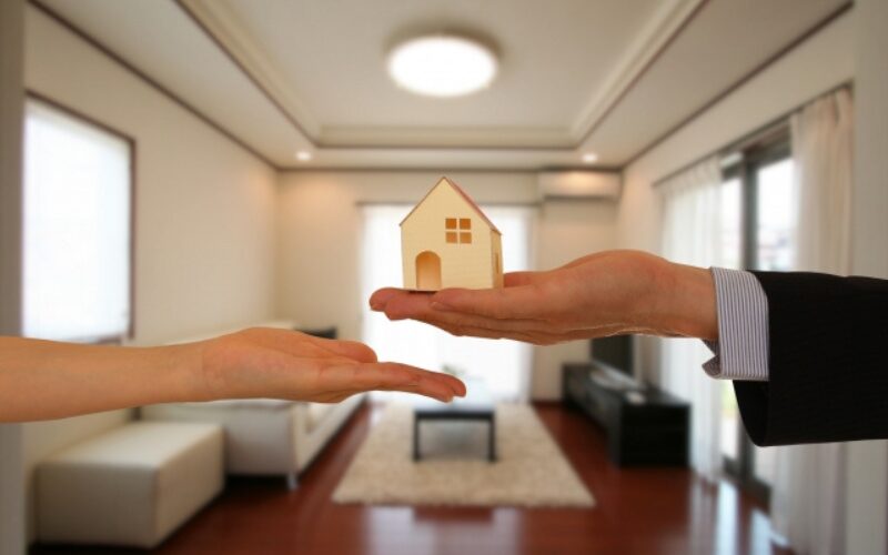 広いリビングルームを背景に、スーツ姿の人物の手から別の人物の開いた手のひらに、小さな家の模型が渡されている