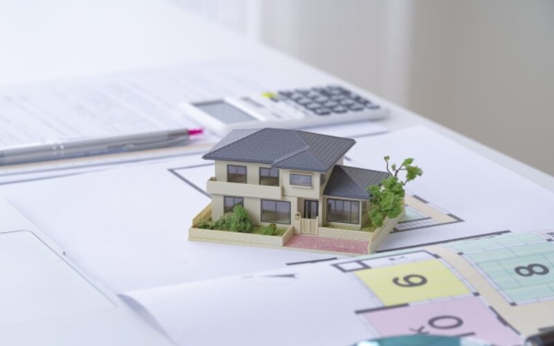 不動産売買契約の流れを検討するために、間取り図の上に置かれた精巧な戸建て住宅の模型