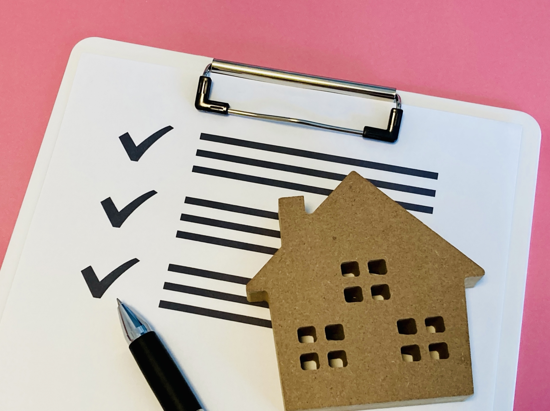 ピンクの背景に置かれたクリップボードのチェックリスト、ペン、小さな木製の家型オブジェ
