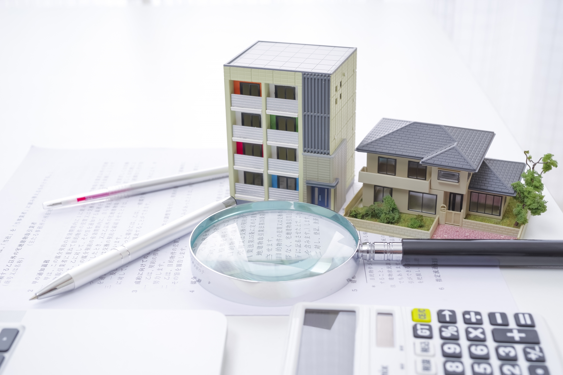 不動産売却査定に必要な書類。マンションと一戸建ての模型、虫眼鏡、電卓