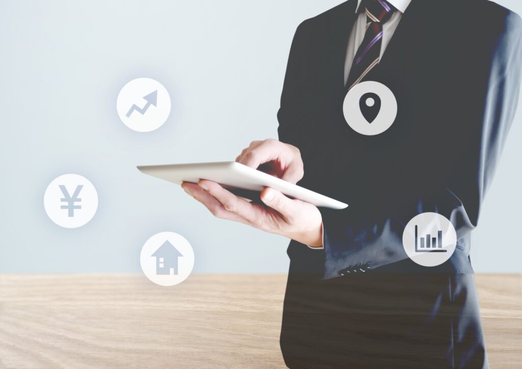 スーツ姿の男性がタブレットを操作しており、周囲に価格、立地、グラフ、家のアイコンが浮かんでいる