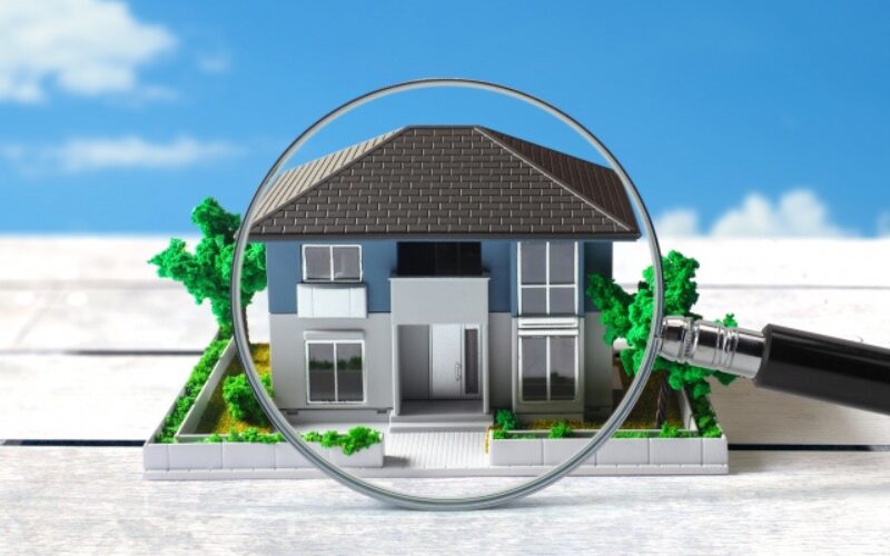 青い空を背景に、青い外壁の二階建て住宅の模型が、虫眼鏡によって大きく拡大されている