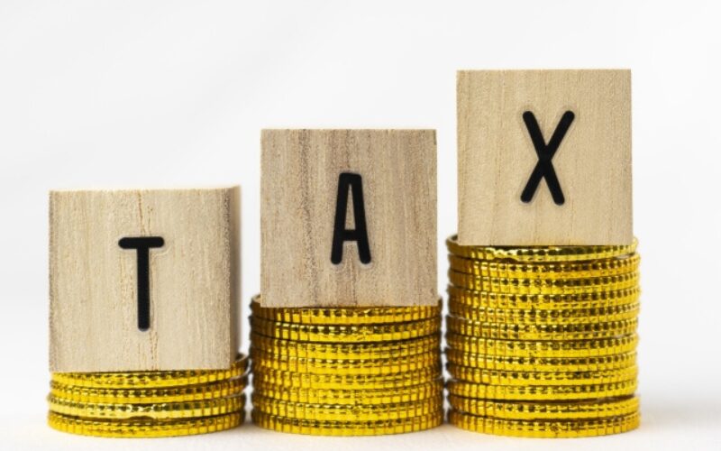 不動産売却で考慮すべき、金色のコインの上に立つ「TAX（税金）」の文字ブロック。