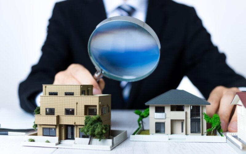 家を高く売るための査定や、売却の判断要素を虫眼鏡で細かくチェックする不動産担当者と戸建ての模型