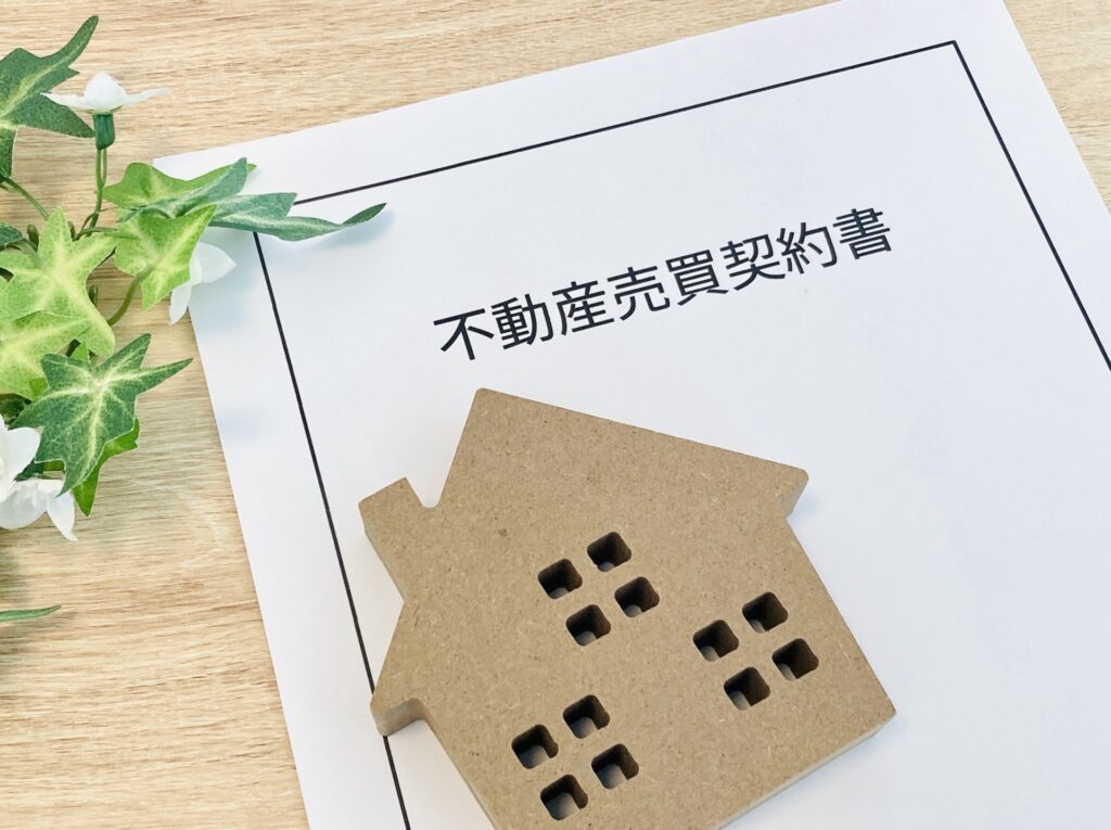 白い書類の上に茶色い家の模型が置かれ、書類には「不動産売買契約書」と黒字で書かれている