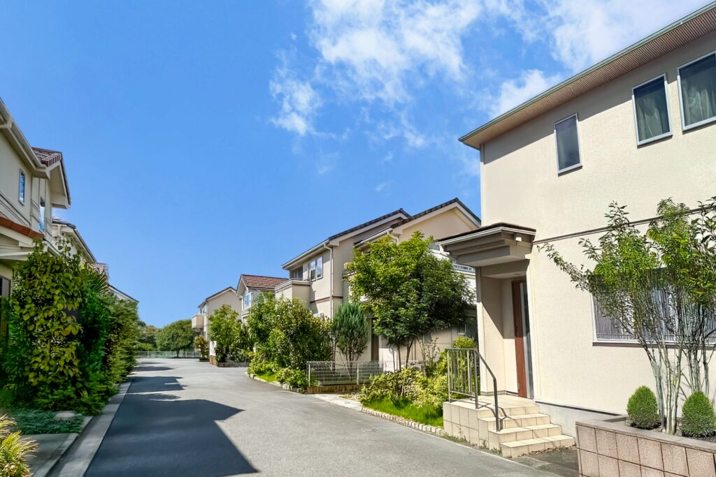 青空の下、並木に沿って続くアスファルトの道と、デザインが統一されたクリーム色の2階建て住宅街の風景。