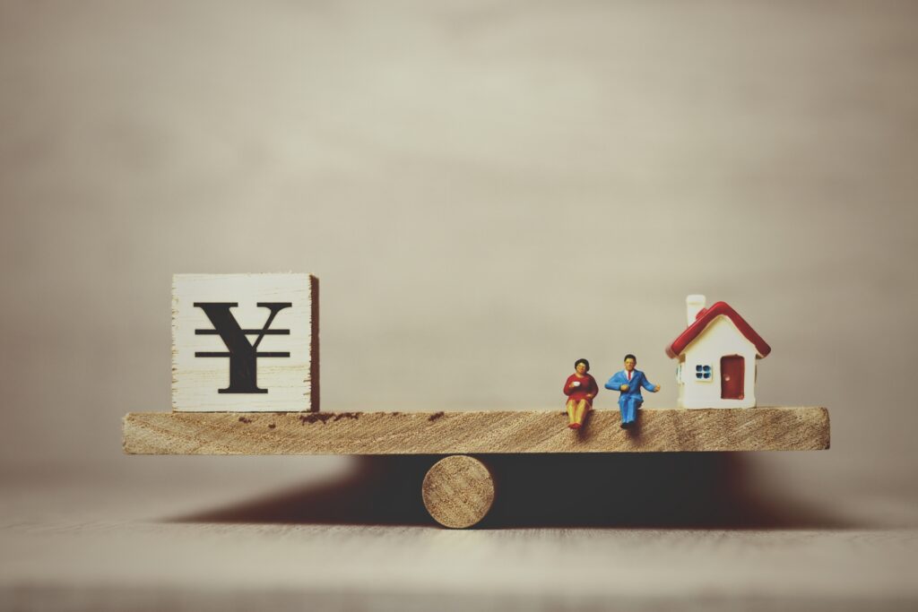 木製のシーソー（天秤）の上に、円マークのブロックと、家の模型、その間に座る二人のミニチュア人形