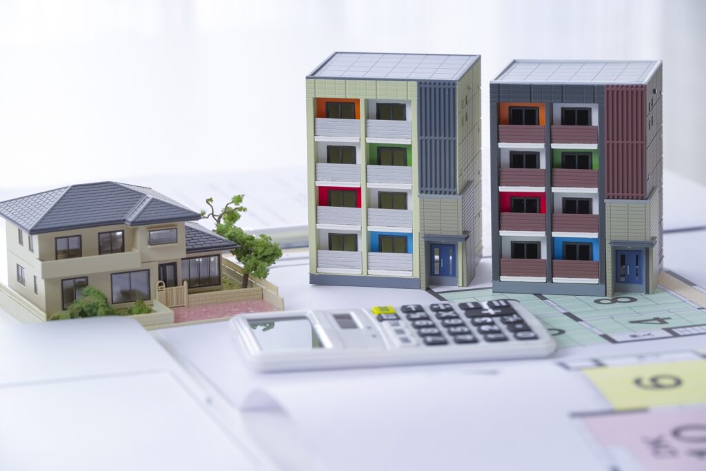 住宅の設計図の上に、一戸建てと2棟のマンション模型、手前に白い電卓が置かれている。